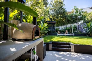 Backyard with a wood-fired pizza oven in the foreground, lush green lawn, and a trampoline and swing set in the background surrounded by tropical plants and trees.